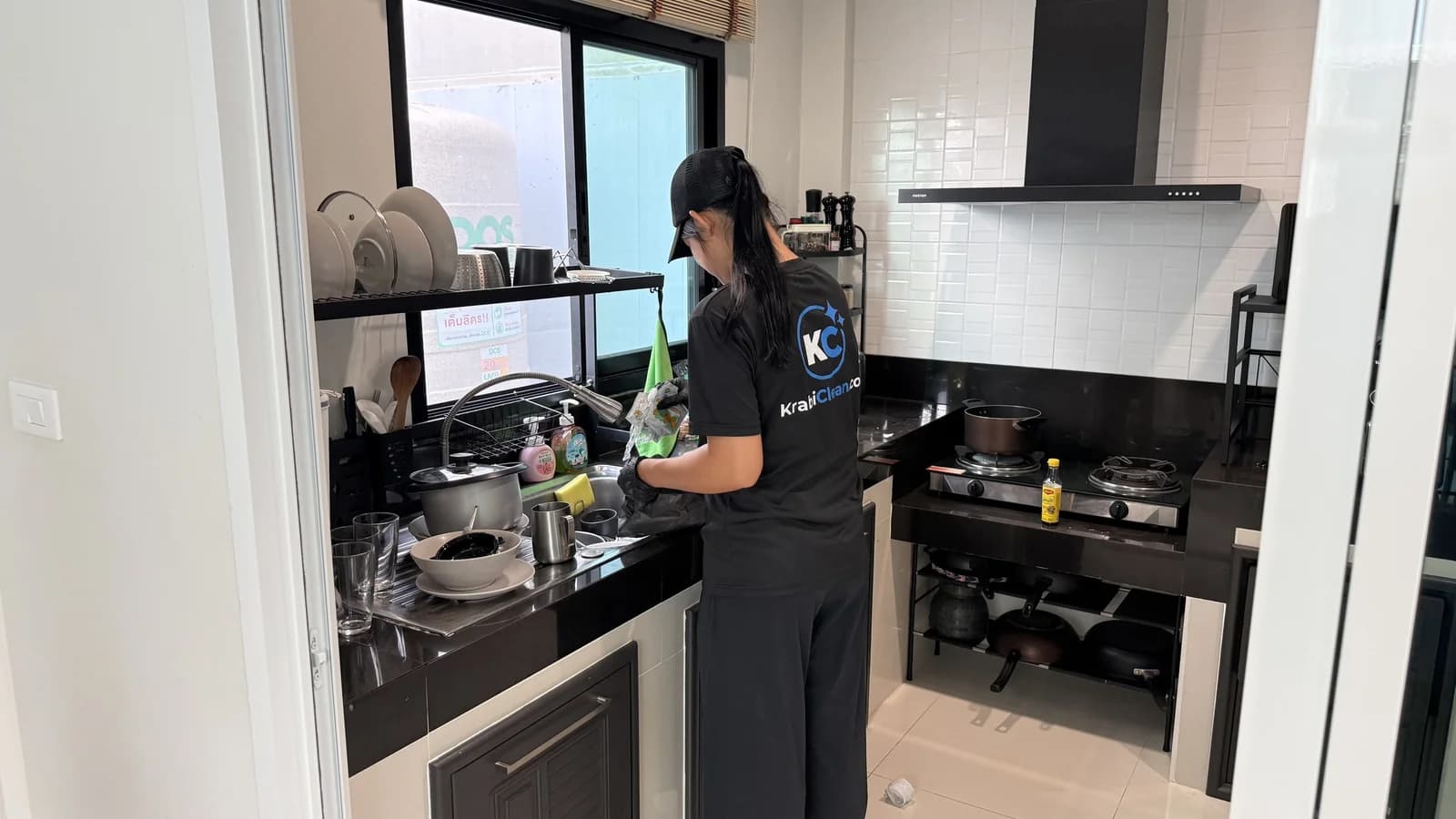 Krabi Clean cleaner in a KrabiClean.com shirt washing dishes at a kitchen sink by the window