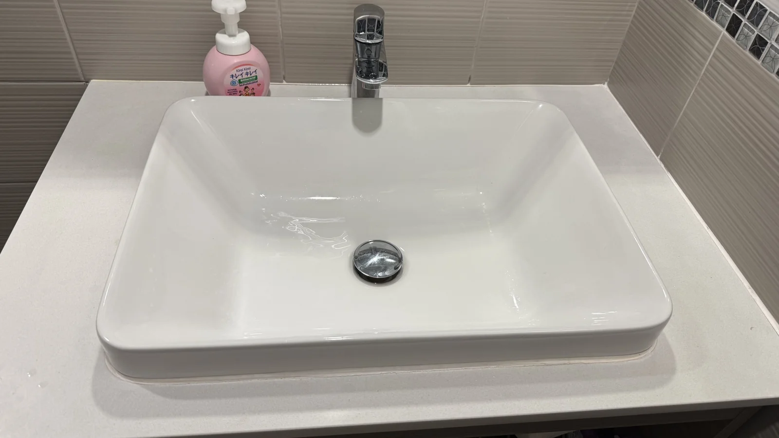 A clean white bathroom basin on a white counter with a chrome tap and a pink soap bottle