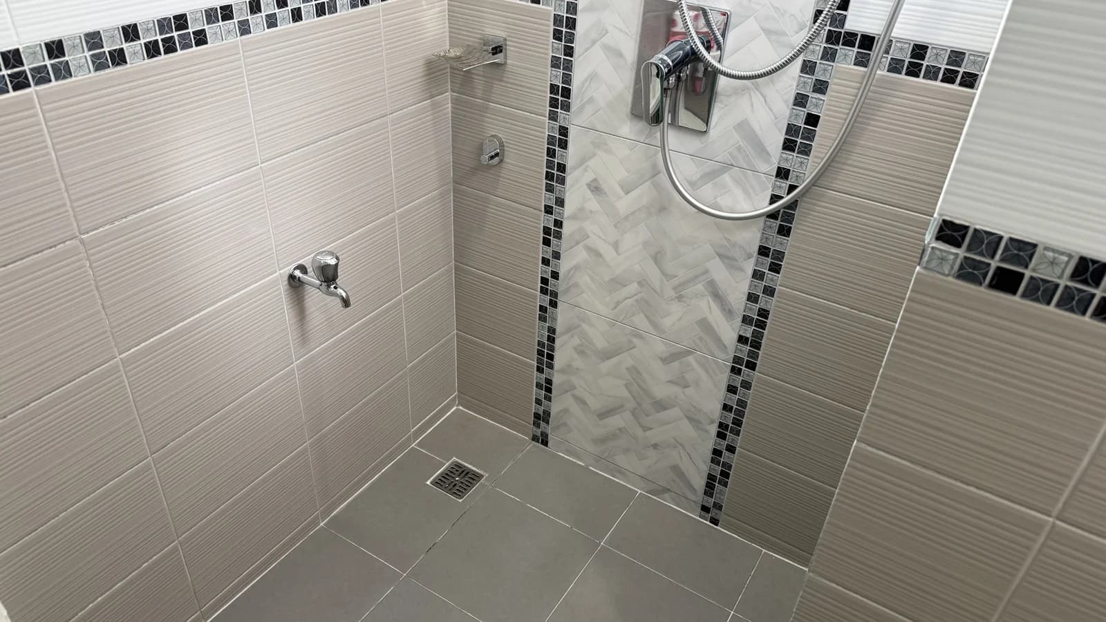 A clean walk-in shower corner with striped wall tiles, a herringbone marble panel, a chrome mixer and a floor drain