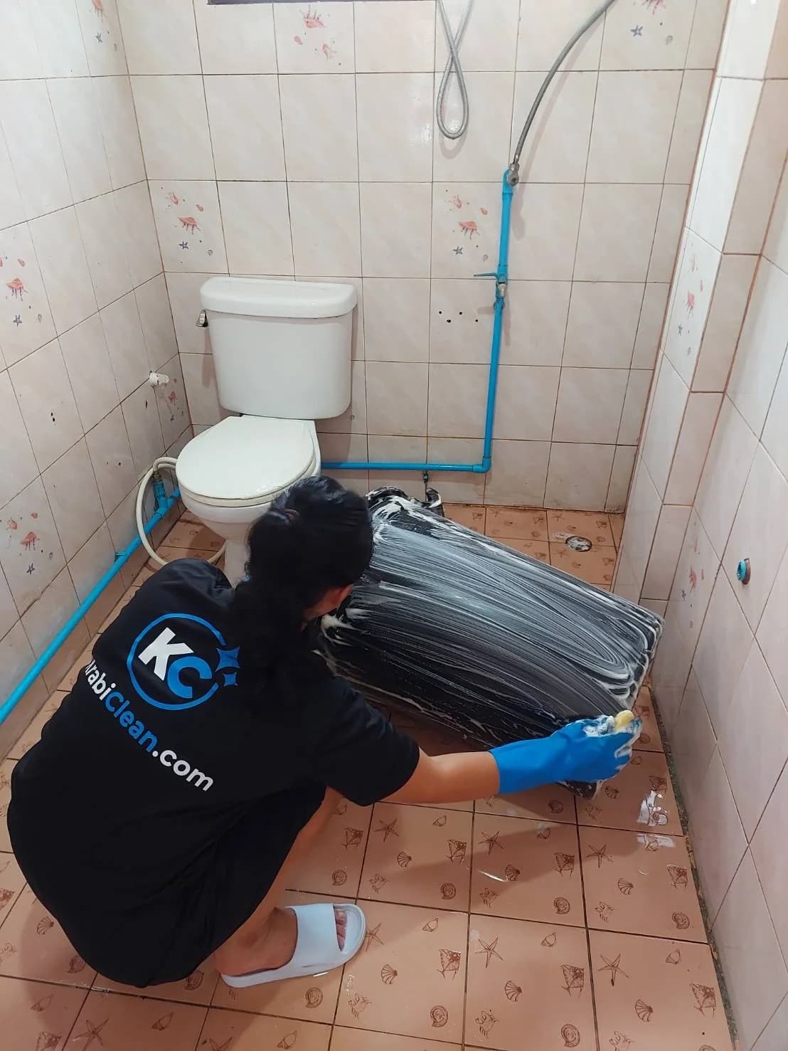 Krabi Clean cleaner in a KrabiClean.com shirt and blue gloves scrubbing a foamy water barrel on a bathroom floor